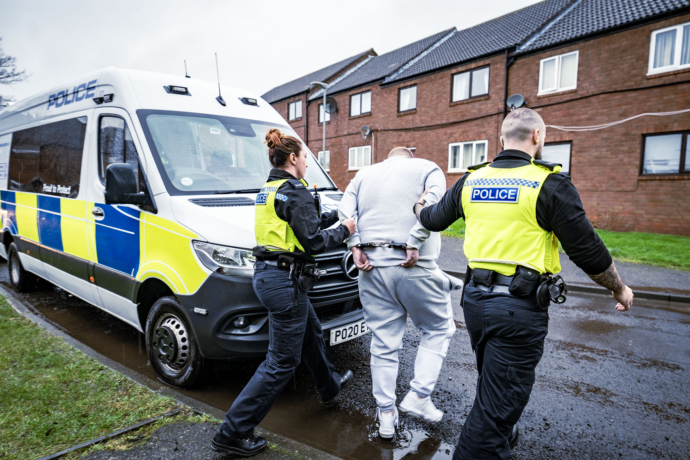 Man in grey being arrested and put into a police van by two police officers
